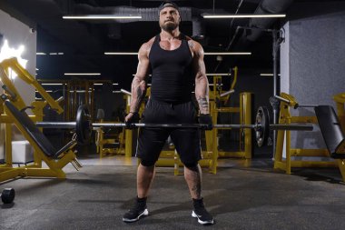 Genç, dövmeli, siyah eldiven, şort, yelek, kep ve spor ayakkabı giyen sakallı bir sporcu. Halter kaldırıyor, kaslarını çalıştırıyor, sarı ekipmanlarla karanlık spor salonunda poz veriyor. Tam uzunluk