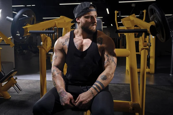Tattooed, bearded athlete man in black sweatpants, vest and cap. He is looking thoughtful while sitting on an exercise machine at dark gym with yellow equipment. Close up