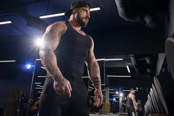 Handsome, tattooed, bearded male in black shorts, vest, cap. Going to do exercises with dumbbells for training his muscles, looking at mirror near set of black weights. Dark gym. Sport. Close up