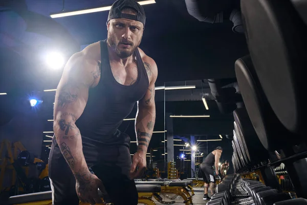 Handsome, tattooed, bearded sportsman in black shorts, vest, cap. Looking at you while standing near set of black dumbbells. Muscular body, dark gym. Fitness, lifestyle. Close up