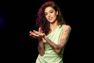 Zarif elbiseli mutlu genç esmer, başarılı poker oyunundan memnun, siyah arka planda duran, bir yığın kazanan bahis fişi tutan. Şans gülümsemesi.