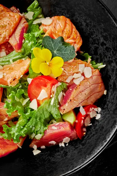 Perfect for dinner, light and refreshing seared tuna steak salad with mixed fresh greens, slices of tomatoes and avocado seasoned with miso dressing garnished with yellow pansy flower in black bowl