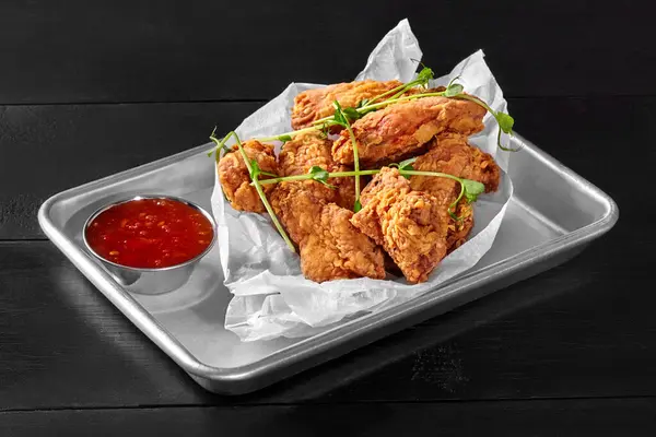 Delicious crispy chicken wings fried in spicy batter presented on baking paper in metal tray accompanied by side of sweet chili sauce, served on rustic black wooden table