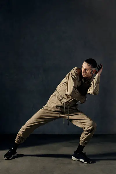 Young handsome performer with tattooed body and face, earrings, beard. Dressed in khaki overalls and black sneakers. He is dancing against gray background. Dancehall, hip-hop. Full length, copy space