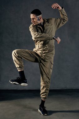 Dövmeli, yüzlü, küpeli, sakallı genç atletik erkek. Haki tulum ve siyah spor ayakkabı giymişti. Gri stüdyo arka planında dans ediyor. Dans salonu, hip-hop. Tam uzunluk, boşluğu kopyala
