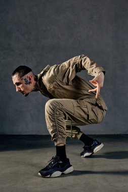 Dövmeli, yüzlü, küpeli, sakallı genç ve çekici bir sanatçı. Haki rengi tulum ve siyah spor ayakkabı giymişti. Gri arka planda dans ediyor. Dans salonu, hip-hop. Tam uzunluk, boşluğu kopyala