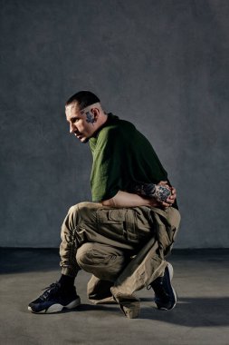 Dövmeli, yüzlü, küpeli, sakallı, esnek bir genç. Yeşil tişört, haki rengi tulum ve siyah spor ayakkabı giymişti. Gri arka planda dans etmek. Dans salonu, hip-hop. Tam uzunluk, boşluğu kopyala