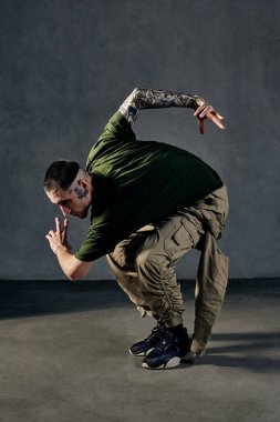 Dövmeli, yüzlü, küpeli, sakallı genç bir atletik. Haki tişört ve tulum giymiş, siyah spor ayakkabı giyiyor. Gri arka planda dans etmek. Dans salonu, hip-hop. Tam uzunluk, boşluğu kopyala