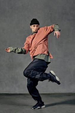 Dövmeli, yüzlü, küpeli, sakallı yakışıklı bir genç. Şapka, günlük kıyafetler ve siyah spor ayakkabılar giymişti. Gri arka planda dans etmek. Dans salonu, hip-hop. Tam uzunluk, boşluğu kopyala