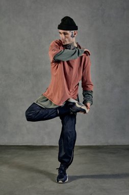 Dövmeli, yüzlü, küpeli, sakallı atletik bir erkek. Renkli kazak, siyah pantolon ve spor ayakkabı giymişti. Gri arka planda dans etmek. Dans salonu, hip-hop. Tam uzunluk, boşluğu kopyala