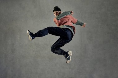 Dövmeli, küpeli, sakallı, atletik bir adam. Şapka, renkli kazak, siyah pantolon ve spor ayakkabı giymişti. Gri arka planda dans ederken zıplamak. Dans salonu, hip-hop. Tam uzunluk, boşluğu kopyala