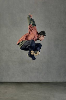 Dövmeli, yüzlü, küpeli, sakallı, görkemli bir genç. Şapka, günlük kıyafetler ve siyah spor ayakkabılar giymişti. Gri arka planda dans ederken zıplamak. Dans salonu, hip-hop. Tam uzunluk, boşluğu kopyala