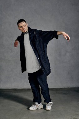 Dövmeli, yüzlü, küpeli, sakallı zarif bir adam. Beyaz tişört, spor ayakkabı, siyah kot gömlek ve pantolon giymişti. Gri arka planda dans etmek. Dans salonu, hip-hop. Tam uzunluk, boşluğu kopyala