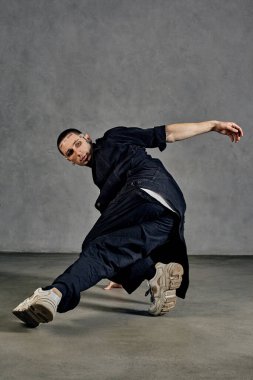 Dövmeli, yüzlü, küpeli, sakallı esnek bir erkek. Beyaz tişört, spor ayakkabı, siyah kot gömlek ve pantolon giymişti. Gri arka planda dans etmek. Dans salonu, hip-hop. Tam uzunluk, boşluğu kopyala