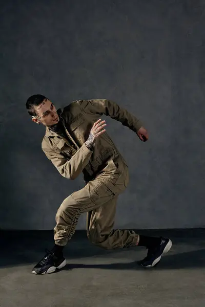 Young graceful male with tattooed body and face, earrings, beard. Dressed in khaki jumpsuit and black sneakers. He dancing against gray studio background. Dancehall, hip-hop. Full length, copy space