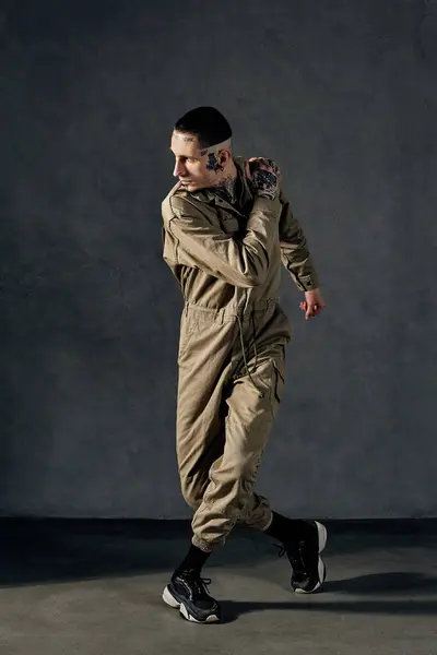 Young graceful fellow with tattooed body and face, earrings, beard. Dressed in khaki jumpsuit and black sneakers. He dancing against gray studio background. Dancehall, hip-hop. Full length, copy space