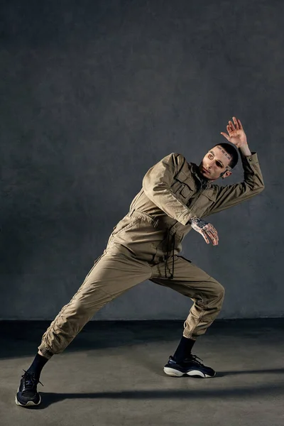Good-looking performer with tattooed body and face, earrings, beard. Dressed in khaki overalls and black sneakers. He is dancing against gray background. Dancehall, hip-hop. Full length, copy space
