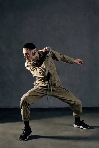 Young stately man with tattooed body and face, earrings, beard. Dressed in khaki overalls and black sneakers. He is dancing against gray studio background. Dancehall, hip-hop. Full length, copy space