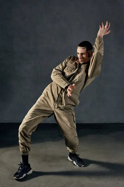 Young stately guy with tattooed body and face, earrings, beard. Dressed in khaki overalls and black sneakers. He is dancing against gray studio background. Dancehall, hip-hop. Full length, copy space