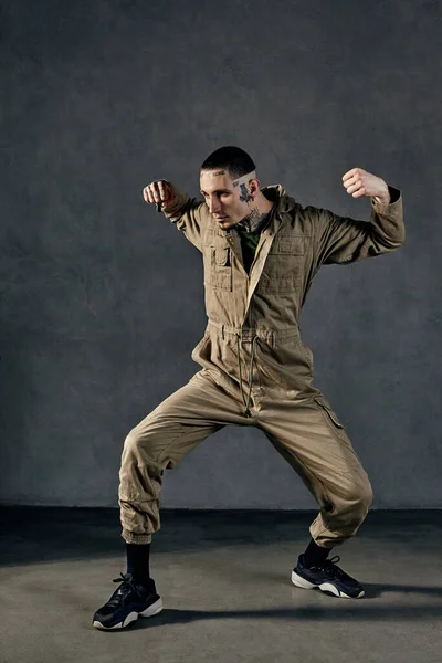 Young active male with tattooed body and face, earrings, beard. Dressed in khaki jumpsuit and black sneakers. He is dancing against gray studio background. Dancehall, hip-hop. Full length, copy space