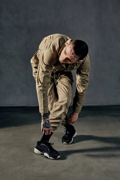 Young flexible male with tattooed body and face, earrings, beard. Dressed in khaki jumpsuit and black sneakers. He dancing against gray studio background. Dancehall, hip-hop. Full length, copy space