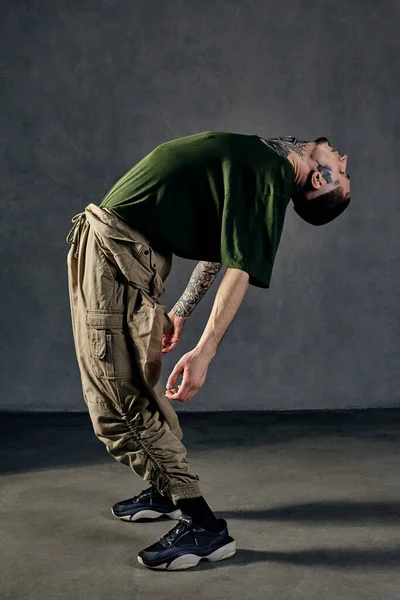 Young graceful guy with tattooed body and face, earrings, beard. Dressed in green t-shirt and khaki overalls, black sneakers. Dancing on gray background. Dancehall, hip-hop. Full length, copy space