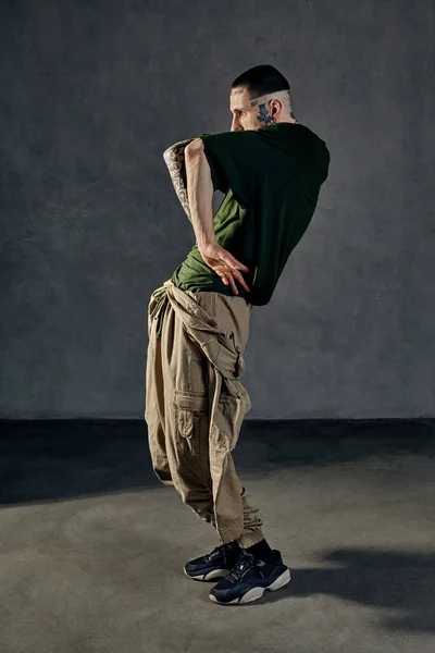 Young athletic man with tattooed body and face, earrings, beard. Dressed in green t-shirt and khaki overalls, black sneakers. Dancing on gray background. Dancehall, hip-hop. Full length, copy space