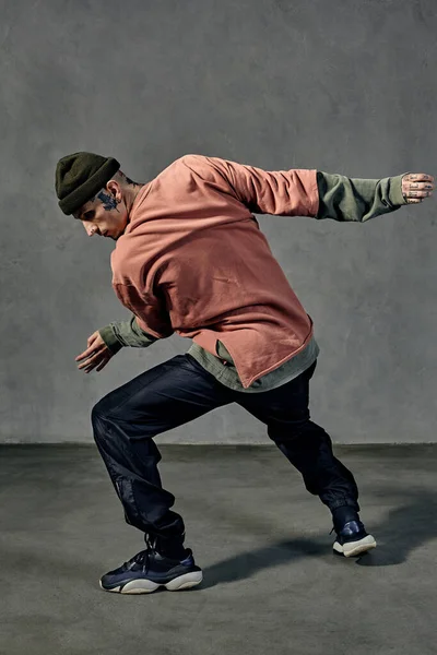 Handsome male with tattooed body and face, earrings, beard. Dressed in hat, colorful jumper, black pants and sneakers. Dancing on gray background. Dancehall, hip-hop. Full length, copy space