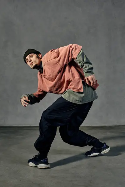 Handsome fellow with tattooed body and face, earrings, beard. Dressed in hat, colorful jumper, black pants and sneakers. Dancing on gray background. Dancehall, hip-hop. Full length, copy space