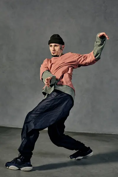 Young graceful male with tattooed body and face, earrings, beard. Dressed in colorful jumper, black pants and sneakers. Dancing on gray background. Dancehall, hip-hop. Full length, copy space