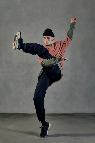 Young athletic guy with tattooed body and face, earrings, beard. Dressed in hat, casual clothes and black sneakers. Dancing on gray background. Dancehall, hip-hop. Full length, copy space