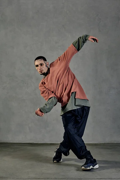 Young stately performer with tattooed body and face, earrings, beard. Dressed in casual clothes and black sneakers. Dancing on gray background. Dancehall, hip-hop. Full length, copy space