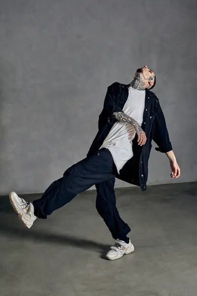 Athletic man with tattooed body, earrings, beard. Dressed in white t-shirt and sneakers, black denim shirt and pants. Dancing on gray background. Dancehall, hip-hop. Full length, copy space