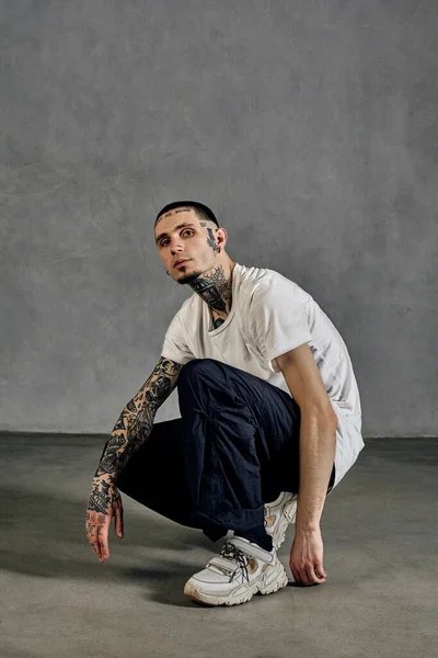 Flexible male with tattooed body and face, earrings, beard. Dressed in white t-shirt and sneakers, black pants. Squatting sideways against gray background. Dancehall, hip-hop. Close up