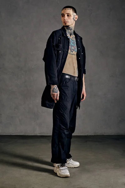Strong guy with tattooed body and face, naked torso, beard. Dressed in black denim shirt, pants and white sneakers. Looking aside, posing on gray background. Dancehall, hip-hop. Full length