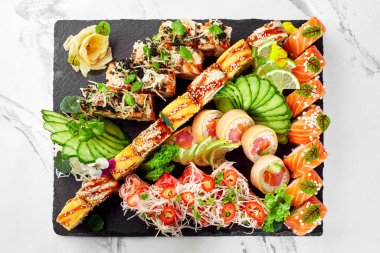 Somon, yılan balığı ve ton balıklı suşi rulolarının müsrif manzarası tahtada sergileniyor ve salatalık, limon dilimleri, çiçekler, acı biber dilimleri ve dilimlenmiş daikon ile süsleniyor.