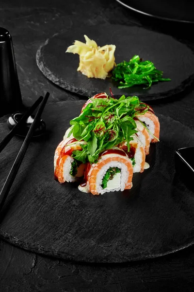Appetizing sushi rolls filled with cream cheese, hiyashi wakame and salmon, topped fresh greens served on black slate board traditionally accompanied by pickled ginger, soy sauce and chopsticks