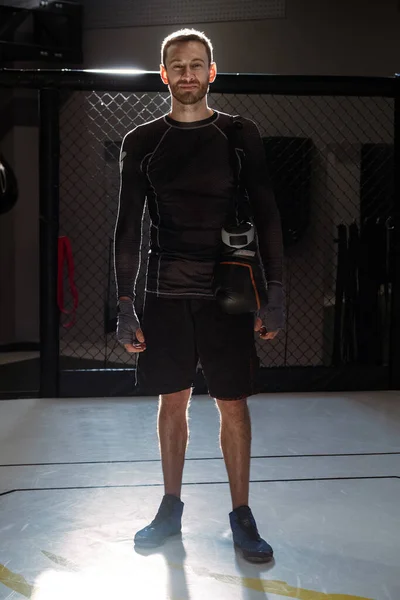 Confident bearded fighter with wrists wrapped in bandages and gloves hanging over shoulder, standing in combat sports cage bathed in sunlight smiling at camera after successful training session