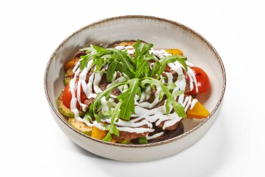 Kırsal seramik kâse iştah açıcı sıcak salata kavrulmuş sebzeler ve kremalı sosla süslenmiş sulu dana eti, taze yeşil roka yapraklarıyla süslenmiş, beyaz arka planda izole edilmiş.