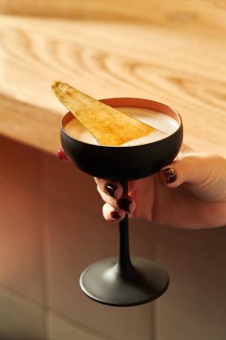 Female hand elegantly holding black stemmed glass with sweetish light alcoholic cocktail garnished with spicy caramelized pear slice against blurred background of wooden bar counter clipart