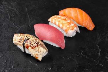 Dört nigiri suşi parçası çeşitli çiğ balık malzemeleriyle siyah bir levha yüzeyinde renkli ve iştah açıcı bir görüntü yaratıyor.