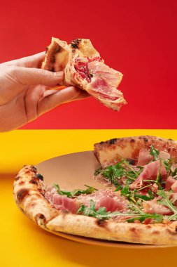 Elleri bir dilim lezzetli pizzayı prosciutto ve roka ile birlikte canlı, renkli arka planda bir tabakta tutar.