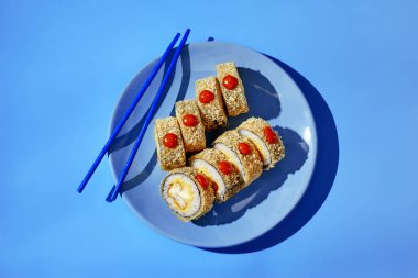 Baharatlı soslu taze tempura maki suşi ruloları mavi yemek çubuklarıyla servis edilir ve canlı bir mutfak sahnesi yaratır.