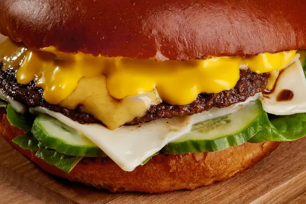 Eritilmiş çedar peyniri, ızgara biftek köftesi, salatalık dilimleri ve kızarmış çöreğin üzerinde marul içeren sulu bir çizburgerin yakın çekimi.