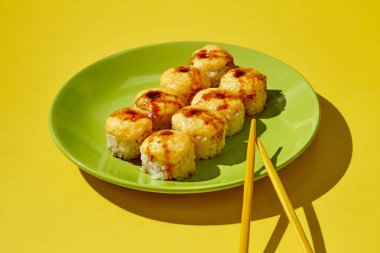 Yeşil tabakta yemek çubuklarıyla lezzetli suşi ruloları, sarı arka planda canlı bir aşçılık sahnesi yaratıyor.