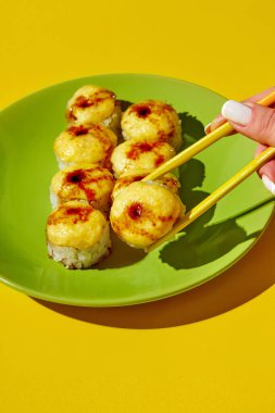 Sarı arka planda yeşil bir tabakta soslu fırında suşi ruloları, yemek çubuklarıyla rulo yapan bir kadın.