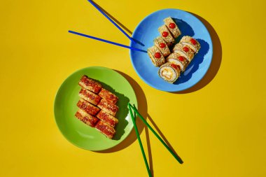 Sıcak kızarmış suşi ruloları ve susam tohumları. Sarı arka planda çubuklarla servis edilen tabaklar yenmeye hazır.