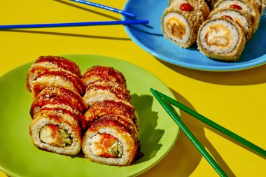 Çeşitli tempura maki suşi rulolarının yakın çekimi renkli tabaklar üzerinde, yemek çubuklarıyla birlikte, görsel olarak çekici bir mutfak sahnesi yaratıyor.