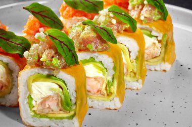 Lezzetli uramaki suşisi peynirli krema, somon balığı, ton balığı ve avakado, yeşil yapraklarla süslenmiş, gri bir tabakta servis ediliyor.