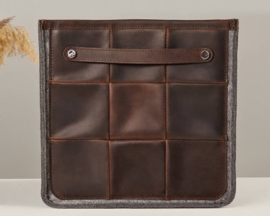 Küçük saplı ve gri keçe kenarlı kahverengi deri çanta tarafsız bir arka planda duruyor.